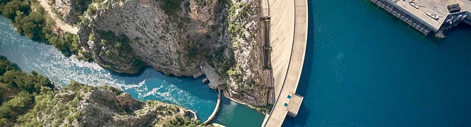 Aerial view of a large curved dam and reservoir. The dam holds back blue water, with a narrow river flowing through rocky terrain nearby.