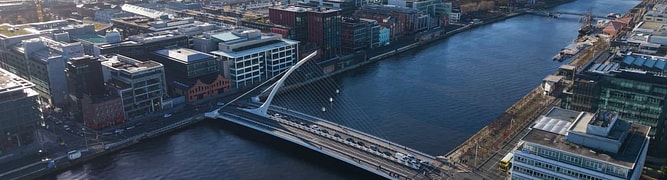Aerial view of a modern bridge spanning a river in a city, with surrounding buildings and roads visible under a clear sky.