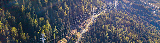 Aerial view of overhead power lines running through a forested mountain landscape under a clear sky, showcasing impressive power line engineering with distant valleys visible.