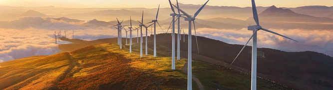 A row of wind turbines stands on a ridge at sunrise, with rolling hills and low clouds in the background.