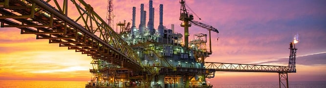 An offshore oil platform stands in the ocean at sunset, showcasing its infrastructure journey with industrial structures, cranes, and a gas flare silhouetted against a colorful sky.