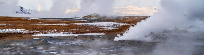 Steam rises from geothermal vents, showcasing the power of geothermal energy in a rocky, snow-dusted landscape under a cloudy sky.