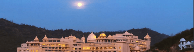 The Fairmont Palace Udaipur hotel pictured at dusk, its intricate Rajasthani architecture illuminated against the hills.