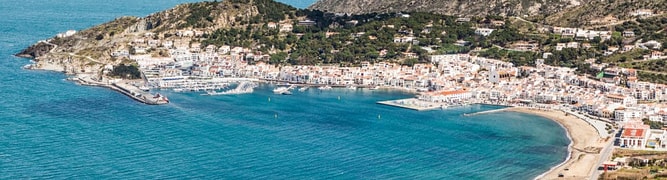 This aerial view features a pale blue sky, deep blue-green ocean, and city with houses featuring white and red roofs built along the bay. Port de la Selva is a traditional fishing village and tourist destination located on the Costa Brava in Catalonia, Spain.