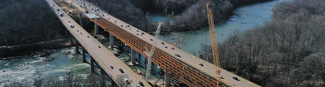 Aerial view of a bridge under construction over a river, with cranes, construction vehicles, and cars on the adjacent completed bridge. 