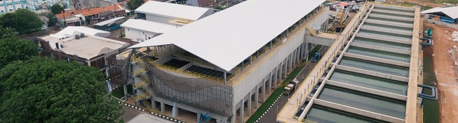 Aerial view of a large industrial facility with white roofing, multiple water tanks, and adjacent buildings in an urban area.