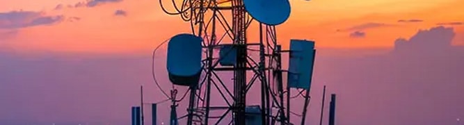 A telecommunications tower with multiple antennas and satellite dishes, overlooking a sprawling cityscape at dusk.