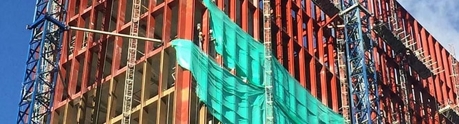 A high-rise research building is under construction with red framework and green safety nets. Blue cranes and scaffolding are visible against a partly cloudy sky, symbolizing a success story in engineering laboratories.