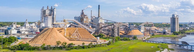 Aerial view of an active coastal pulp and paper mill featuring processing towers, smokestacks, and large wood chip piles which was reconfigured by Delta Wye Engineering using EasyPower.