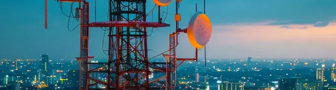 A telecommunications tower with multiple antennas and dishes overlooks an illuminated cityscape at dusk. 
