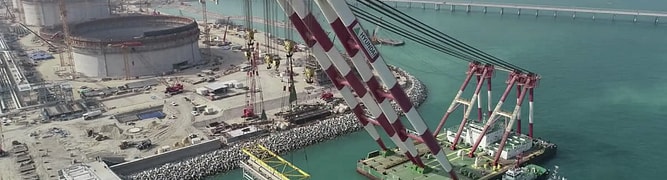 A large crane barge lifts precast concrete structures near storage tanks and a construction site at a waterfront industrial facility. Tugboats assist in the operation.