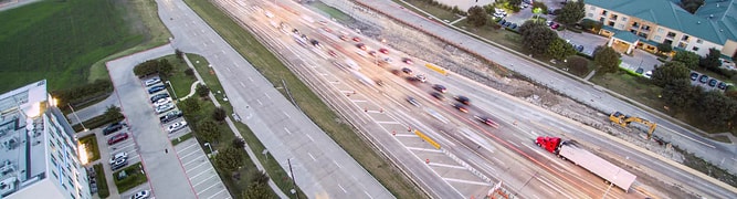 An aerial view of a busy highway in Plano, Texas, with blurred evening traffic flowing past the commercial district.