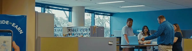 Four people stand and talk around a tall desk in a modern office with cubicles and large windows. A poster with a phone is visible on the left, highlighting the latest site design solutions like Bentley OpenSite+.