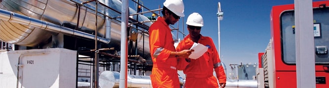 Two engineers in safety gear review documents on-site at Cairn India's Mangala Processing Terminal in Rajasthan, with large, insulated pipes and vessels in the background.