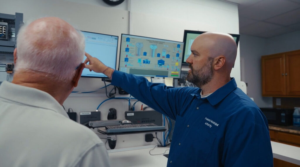 Two men in a lab with the Freeland Municipal Authority review data on wall-mounted computer screens; one man gestures toward a monitor while explaining how digital twins can help address water loss.