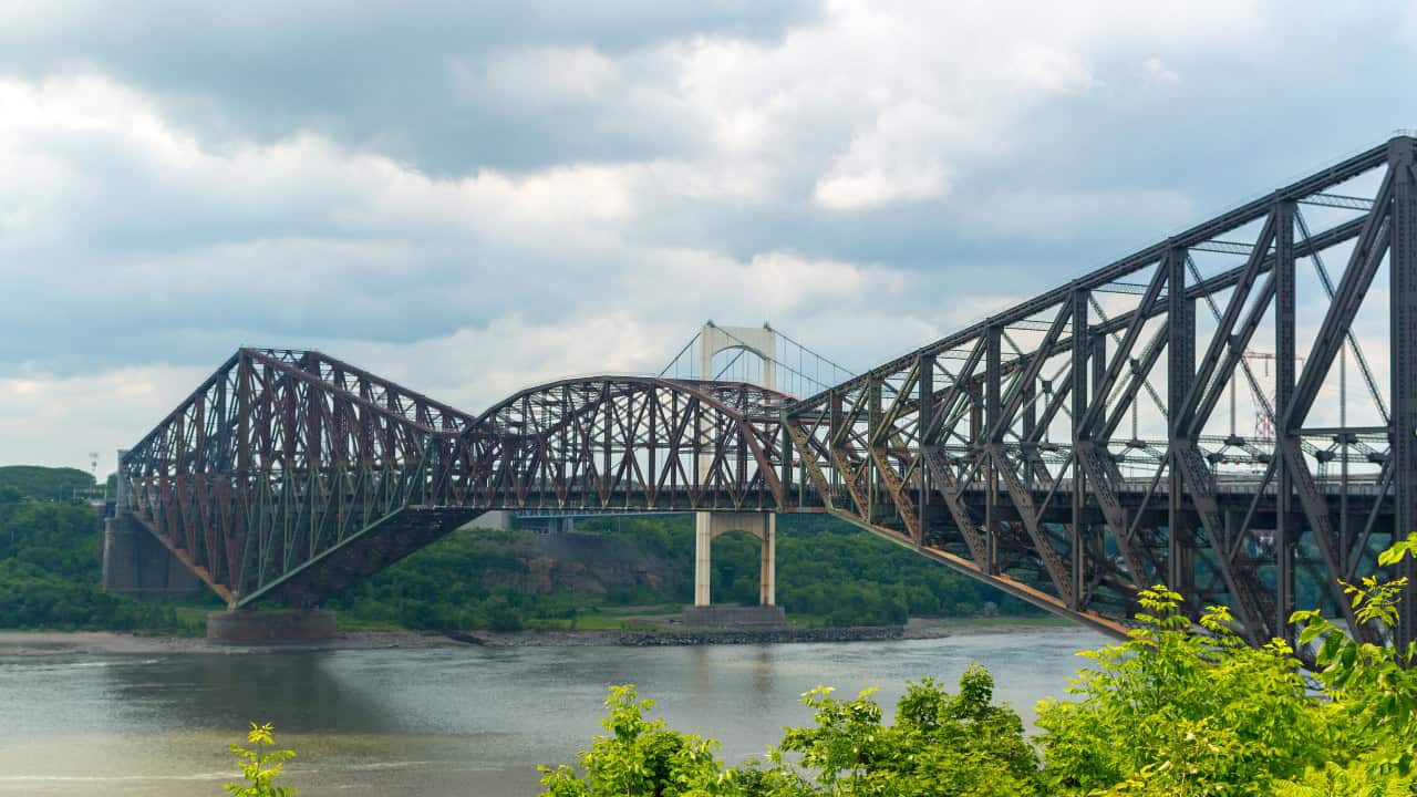 Large steel truss bridge spans a river under a cloudy sky, with another bridge visible in the background and green foliage in the foreground&mdash;a true engineering wonder reminiscent of the iconic Quebec Bridge.