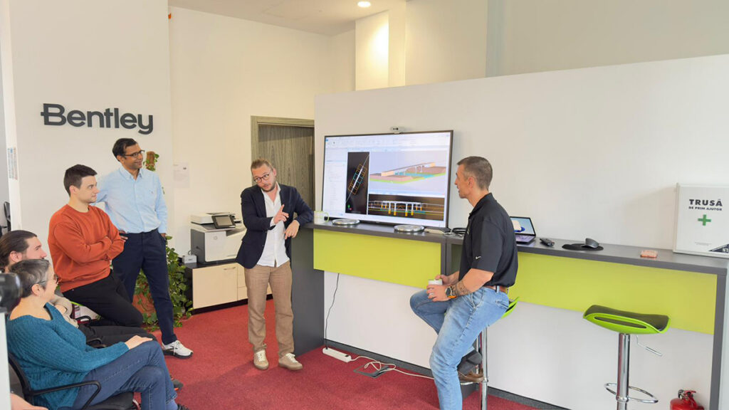 Group of professionals listening to a presenter in a Bentley office, a screen shows architectural CAD designs on a large monitor next to the Bentley sign.