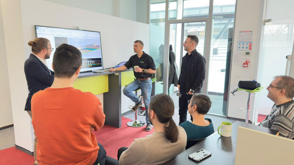 Six people are gathered in an office, looking at a large monitor displaying a design or model created with Bentley’s OpenBridge Designer. Some are seated, others are standing, and a man is presenting with a mug in hand, reinforcing the software trust that brings their collaborative project to life.