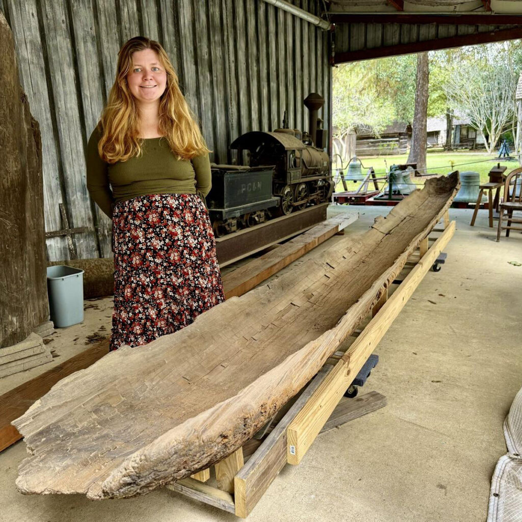 A woman stands next to a large, weathered wooden canoe displayed on a wooden support structure in a covered outdoor area, evoking reflections on the history of travel and exploration—contrasting with today’s innovative visions for playable cities and future space habitats.