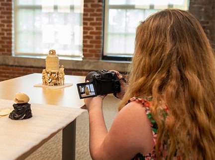 A person with long hair uses a camera to photograph ivory artifacts displayed on a table in a well-lit room with brick walls and large windows, evoking the feeling of exploring playable cities or documenting historical pieces for future digital twin technology archives.