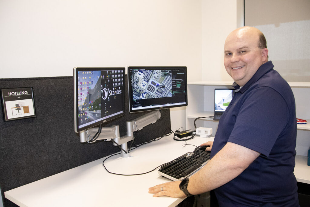 A man in a blue shirt smiles at a standing desk with two monitors displaying maps and software in an office workspace labeled "Hoteling," addressing staffing challenges with the help of AI in infrastructure solutions.