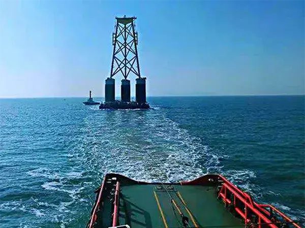 A barge tows a large metal wind turbine foundation with cylindrical floats across a calm sea under a clear blue sky.
