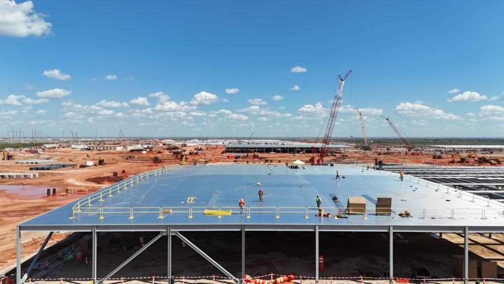 A large construction site in Texas with workers on a steel platform, cranes, and materials spread across a future hyperscale AI data center, built by DPR Construction under a blue sky with scattered clouds.