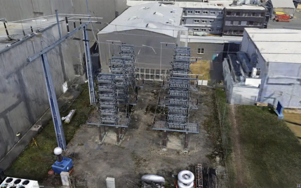 A fenced area with electrical transformers and insulators is situated next to several industrial buildings, with overhead power lines running through the site.