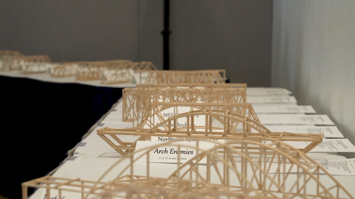 Wooden lattice bridge models displayed on a long table with project name cards visible at the front.