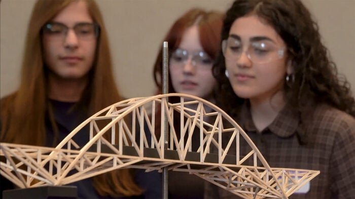 Three students wearing safety glasses observe a scale model of a wooden truss bridge placed on a table indoors.