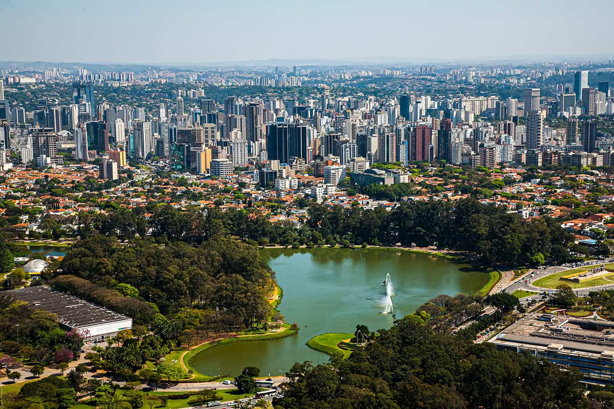 Aerial view of a cityscape with a large park, a lake with a Smart Water fountain by Sabesp, trees, and high-rise buildings in the background under a clear sky.