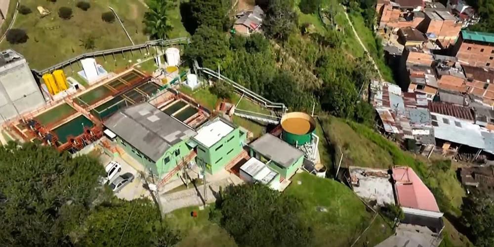 Aerial view of an EPM water treatment facility in Medellín, with green buildings and tanks providing reliable water next to a densely built residential neighborhood and lush vegetation.