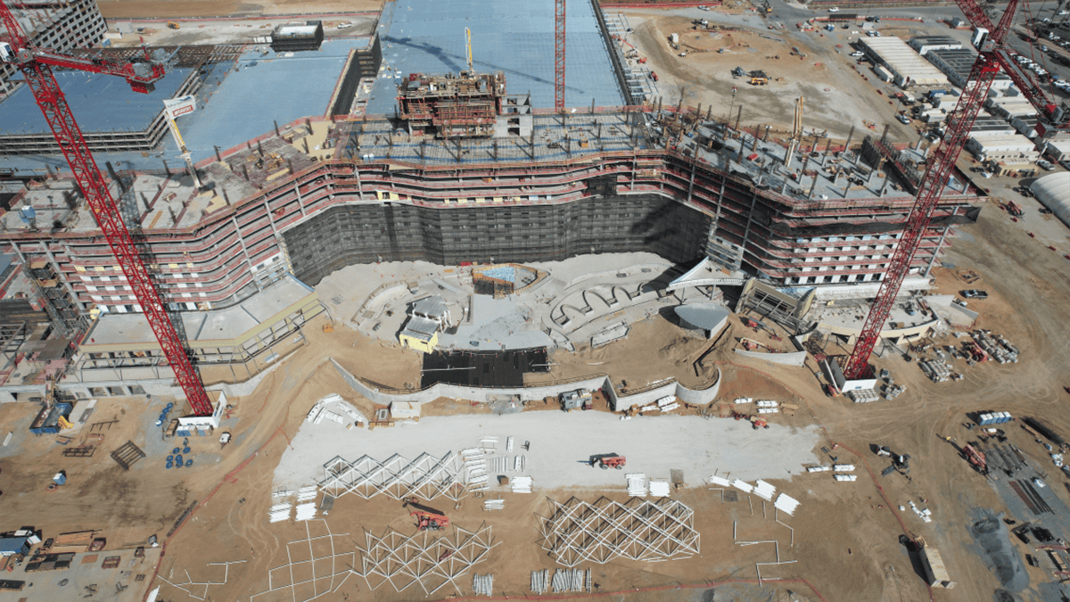 Aerial view of a large construction site with cranes, partially built structures, and building materials spread across sandy ground.