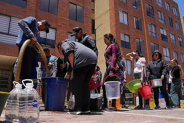 People fill containers with water from a hose on a city sidewalk, standing in line with various buckets and jugs, each waiting for their turn to access this vital resource—a reminder of the power water holds over life on our planet.