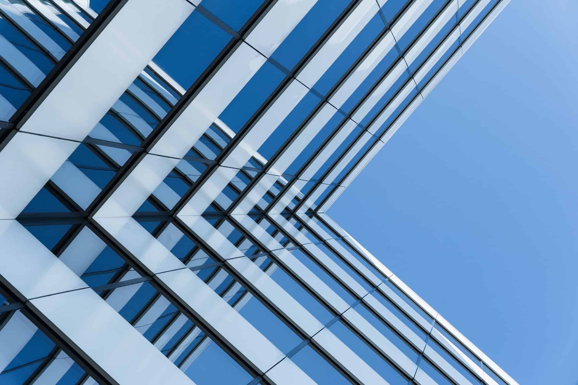 A modern glass office building with reflective windows viewed from below, showing geometric lines against a clear blue sky.