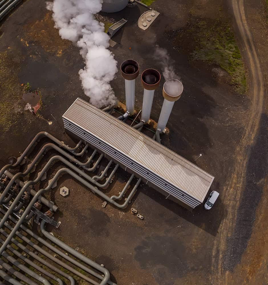 Aerial view of an industrial facility with three large smokestacks emitting steam or smoke and multiple pipelines on a dark, barren ground, showcasing how digital transformation can enhance asset performance.