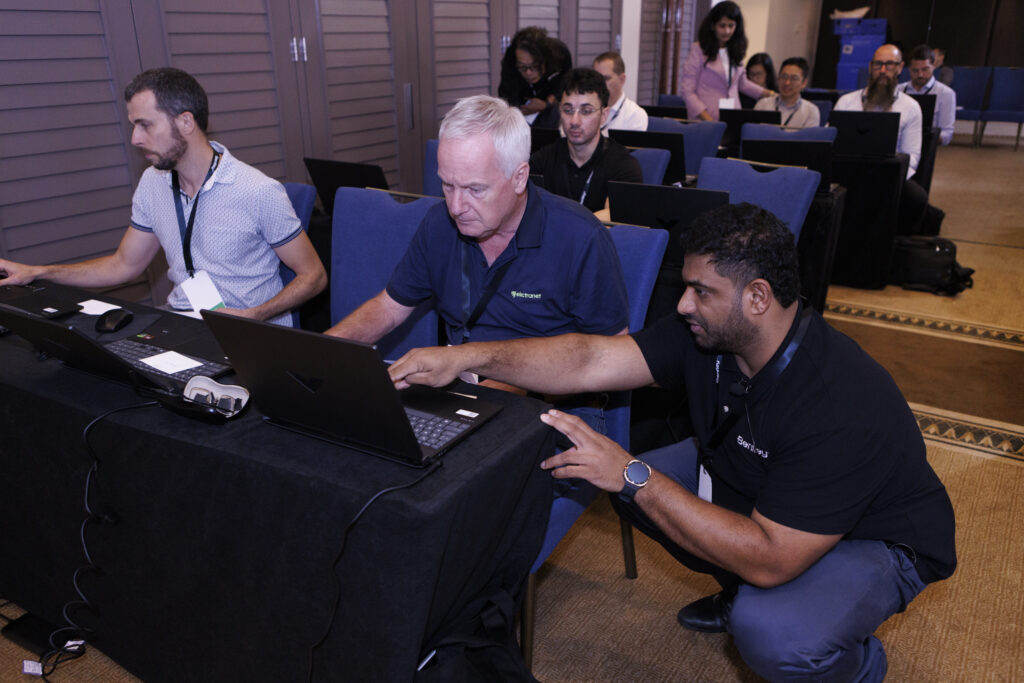 A man kneels next to a seated man, assisting him with a laptop during a workshop or training session with other participants at tables in the background.