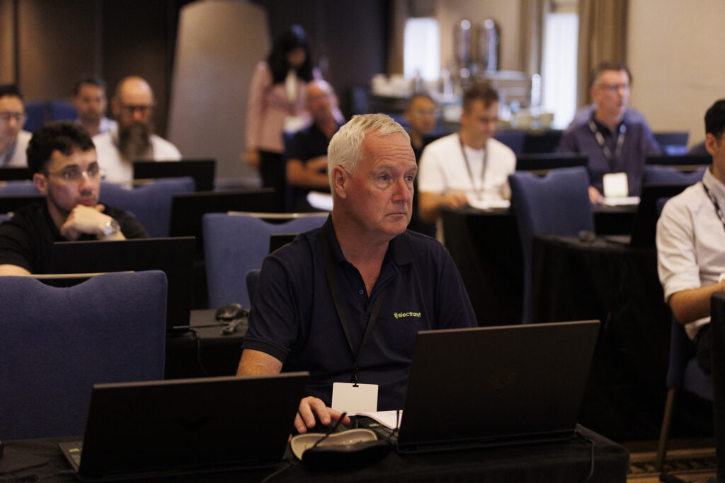 A group of people seated in a conference room, with laptops open; a man in the foreground is focused on his screen.