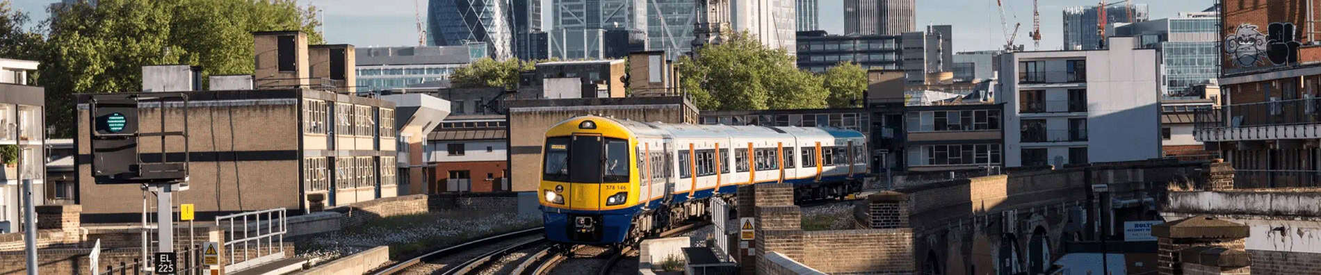 A modern yellow and blue train, a testament to engineering clarity, travels on elevated tracks through an urban area with buildings and skyscrapers in the background.