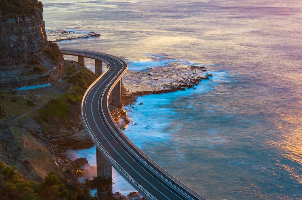 A curved coastal bridge, envisioned by civil engineers, extends over the ocean and connects rocky cliffs with the sea below during a sunset or sunrise.