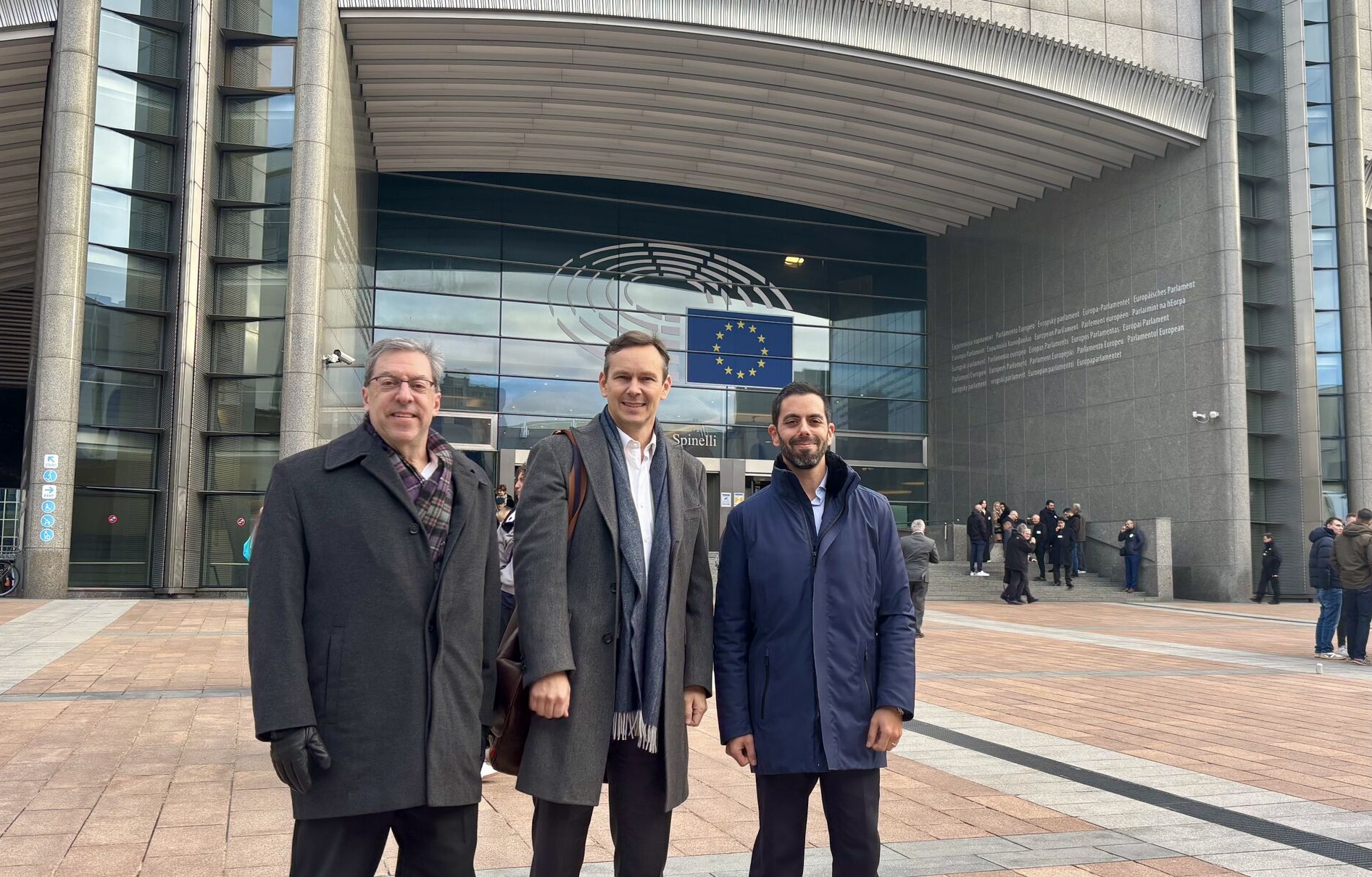 Three men in business attire stand outside the entrance of a large modern building with the European Union flag displayed above.