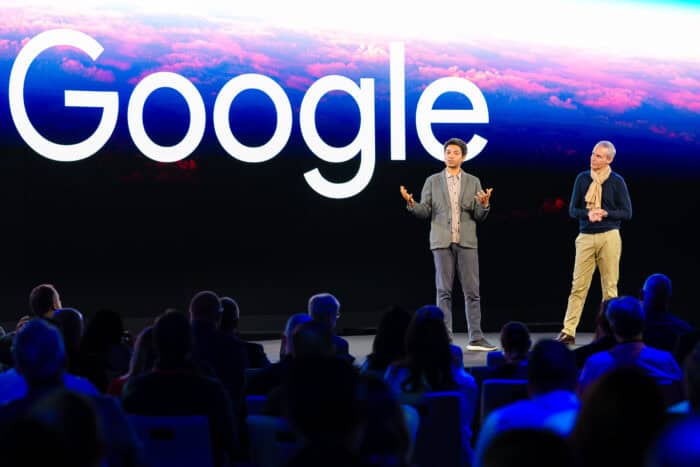 Two people stand on stage in front of a large screen displaying the word Google, addressing an audience.