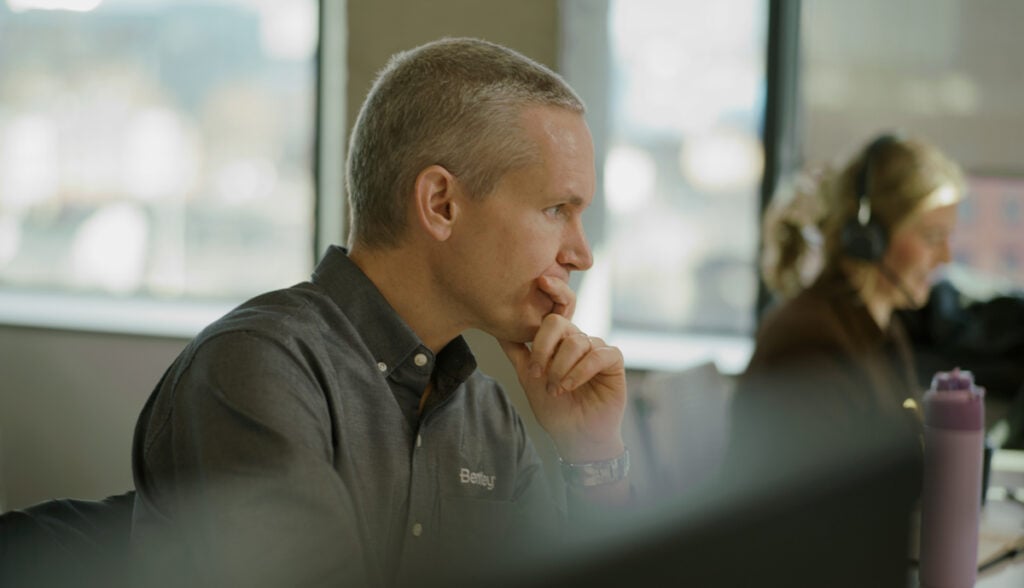 A man with short gray hair sits at a desk, looking at a computer monitor with his hand on his chin. Another person with headphones is in the background.