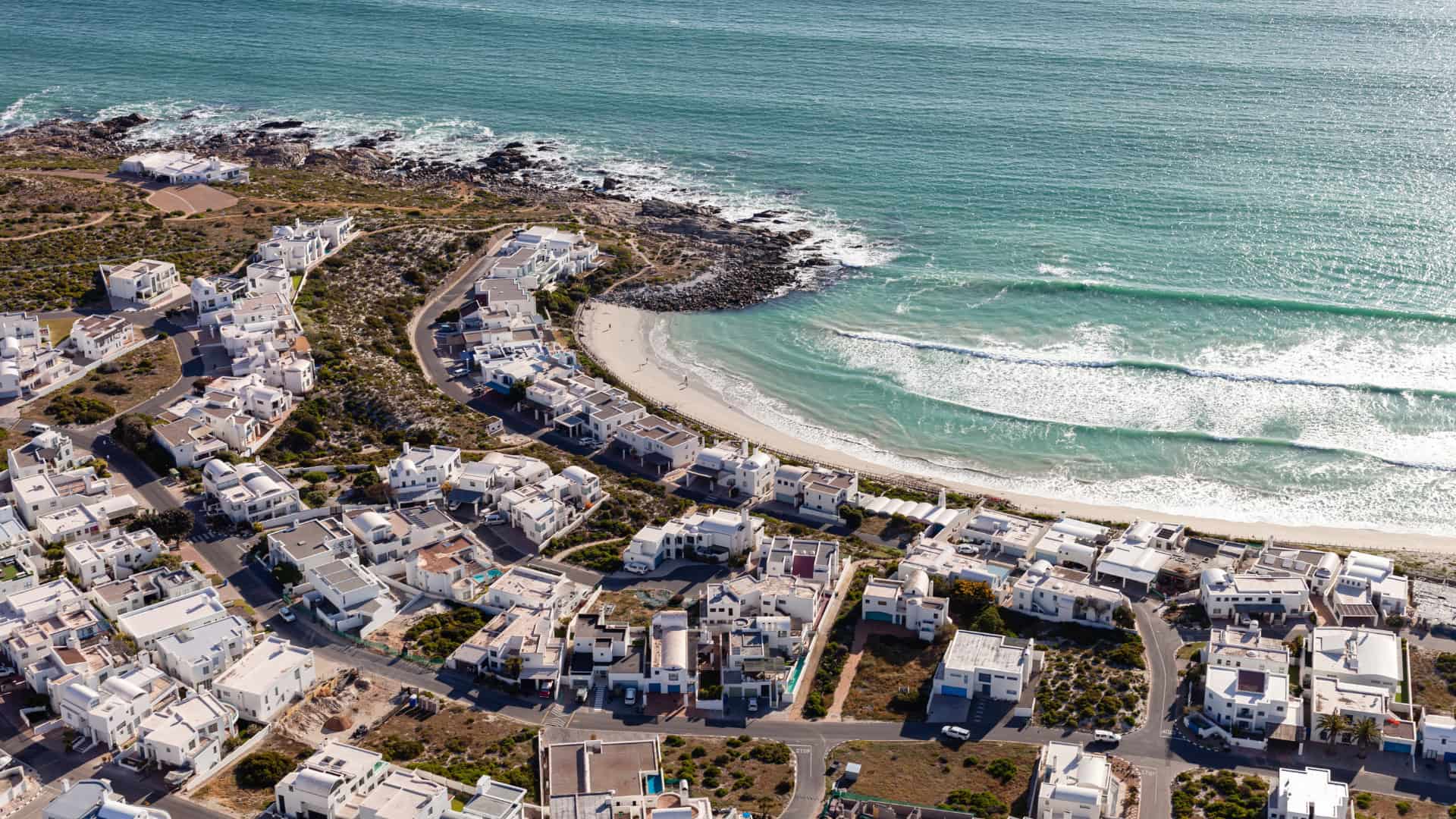 Aerial view of a coastal South Africa town with white houses, curved streets, and a sandy beach by turquoise ocean waves shaped by climate change.