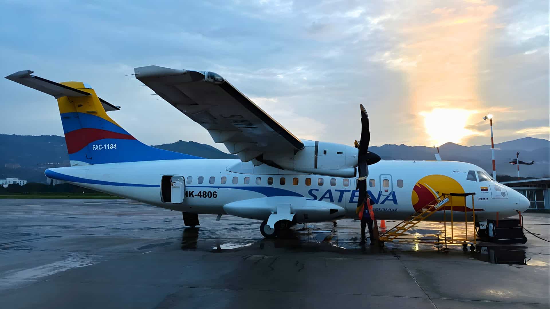A SATENA ATR 42 aircraft is parked on a wet tarmac at sunset in Colombia, with boarding stairs at the front door and mountains in the background, ready to keep isolated towns connected across the region.