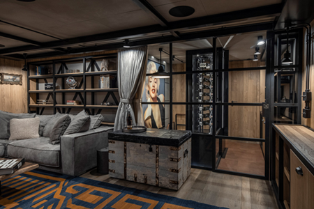 An image of a modern industrial-style room features a gray sofa, shelves, a large trunk, and a glass enclosure with a large portrait of a woman.