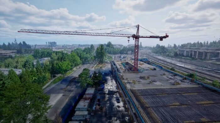 A large red construction crane stands over a building site next to a road and railway tracks, surrounded by trees and several buildings under a partly cloudy sky.
