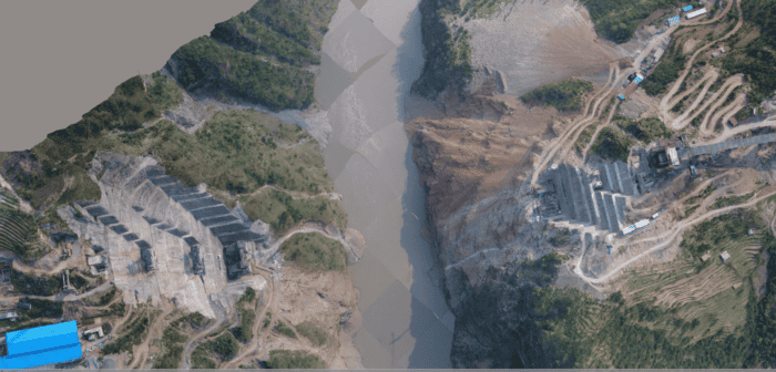 Aerial view of a river separating two construction sites surrounded by green vegetation. The left side has several structures including a blue-roofed building, and the right side shows ongoing earthworks.