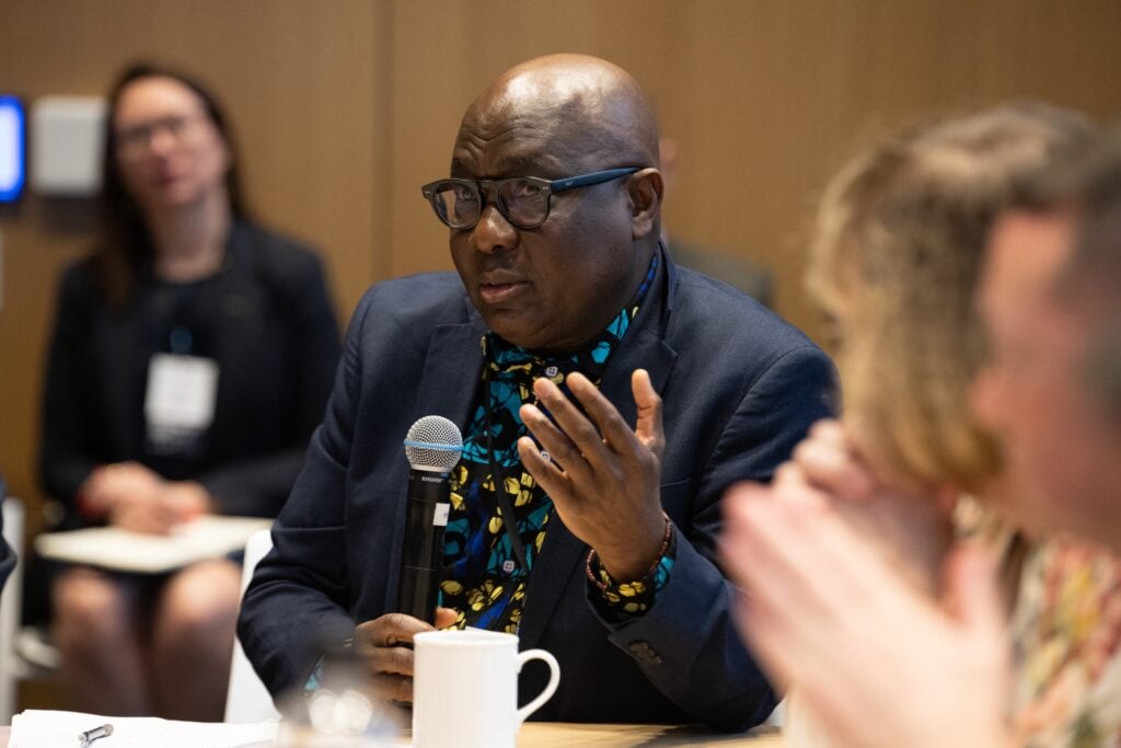 A man in glasses speaks into a microphone during a meeting on U.S. infrastructure, with other participants gathered around the conference table, discussing actionable data and AI-driven solutions.