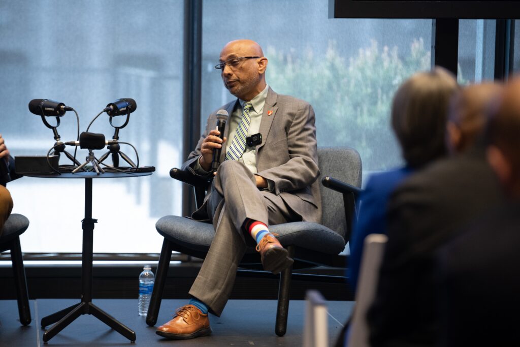 A man in a suit sits on a chair with legs crossed, speaking at an indoor panel event on U.S. modernization, with microphones and actionable data materials on the table beside him.